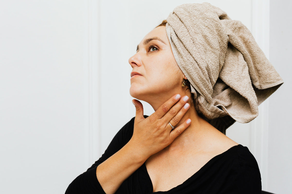 Woman with a towel wrapped around her hair, using her hands to apply a product to her neck