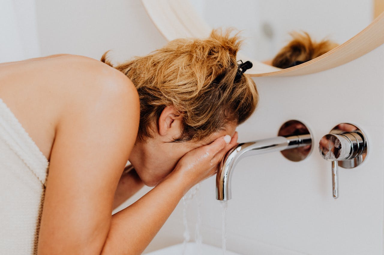 woman wrapped in a white towel bending over a sink, washing her face