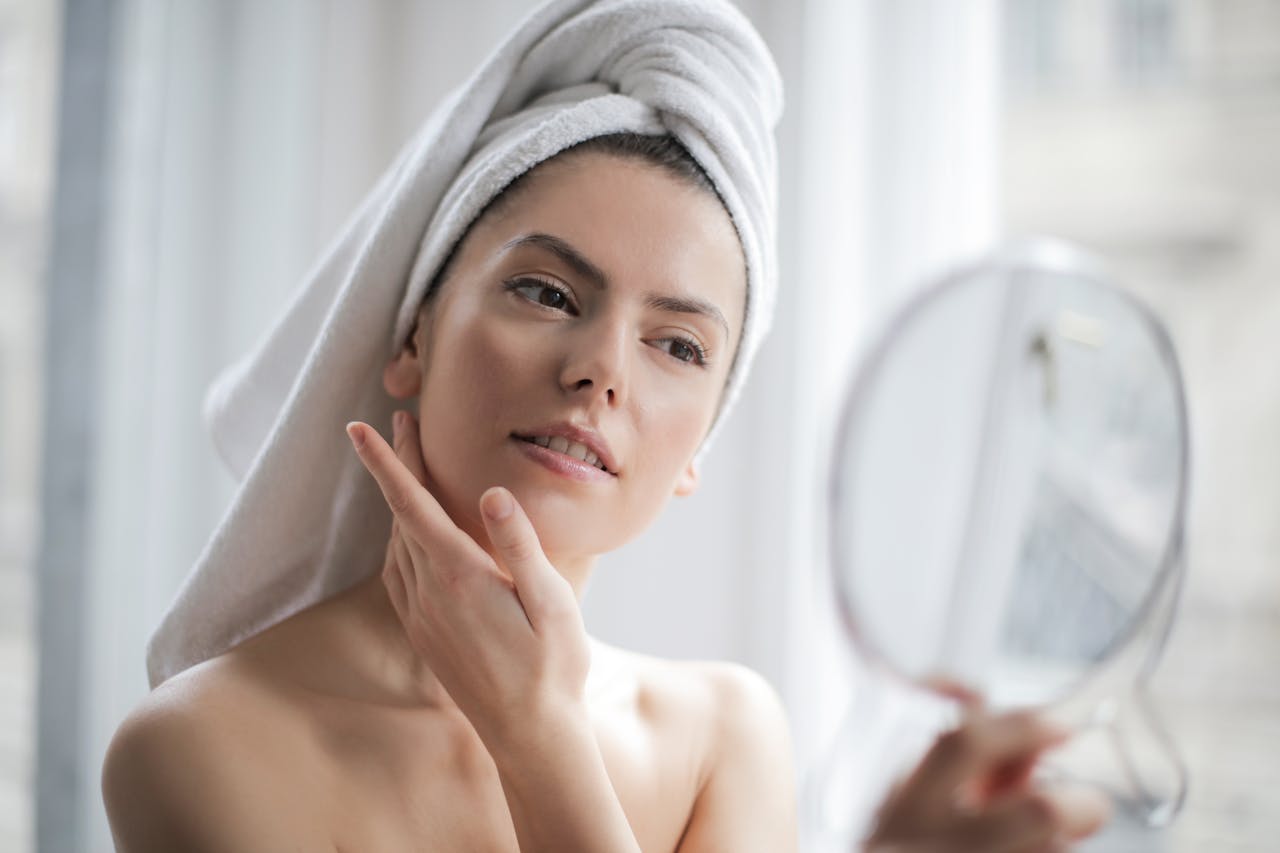 woman with her hair wrapped in a towel looking into a mirror with her hand touching her face